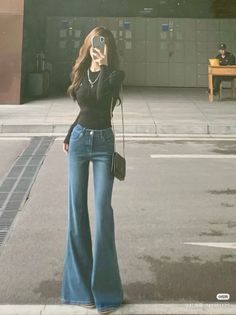 Woman in a black top and bell bottom jeans taking a mirror selfie outdoors
