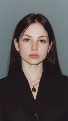 Woman with long dark hair wearing a black jacket and necklace against a plain background