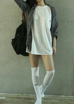 Person in a plaid shirt, white t-shirt, and knee-high socks against a concrete wall