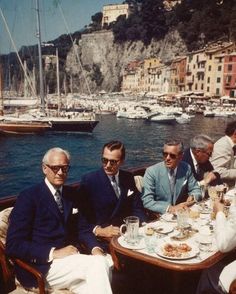 Men in suits and sunglasses dining on a boat in a harbor with a cityscape background