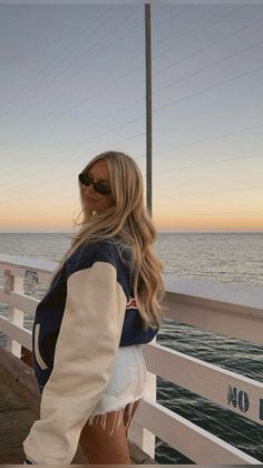 Woman with long blonde hair wearing a varsity jacket and denim shorts on a pier at sunset