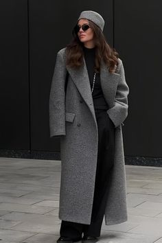 Woman in a long gray coat, black pants, and a hat standing against a dark background