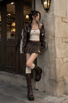 Woman in a brown leather jacket and plaid skirt leaning against a stone wall