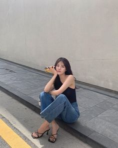 Woman with long dark hair sitting on a curb in a black top and blue jeans