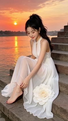 Woman in a white dress sitting on steps by a river at sunset