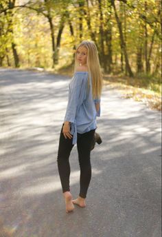 Young woman with long blonde hair in an off-shoulder top and black pants, barefoot on a road