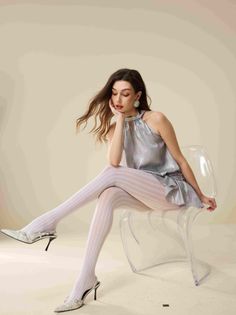 Woman with long hair wearing a silver top and white tights sitting on a clear chair