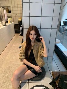 Woman with long hair wearing a tan jacket and black dress, sitting indoors