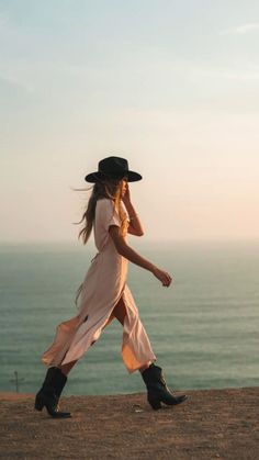 Woman in a long dress and hat walking along a cliff overlooking the ocean at golden hour