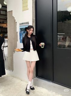 Woman in a black cardigan and white pleated skirt holding a drink and pastry