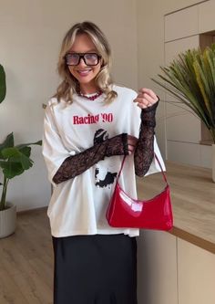 Blonde woman in glasses, white t-shirt, and black skirt holding a red handbag