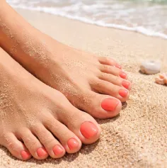 Feet with coral nail polish on a sandy beach with ocean waves