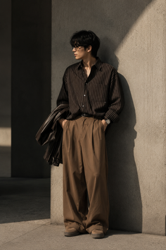 Man in a striped shirt and wide leg pants leaning against a wall outdoors