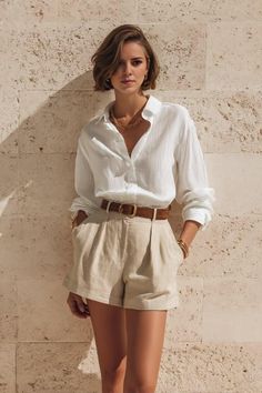 Woman with short hair wearing a white shirt and linen shorts against a stone wall