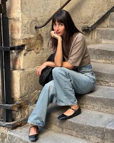 Woman with long hair and bangs sitting on stone stairs in jeans and ballet flats