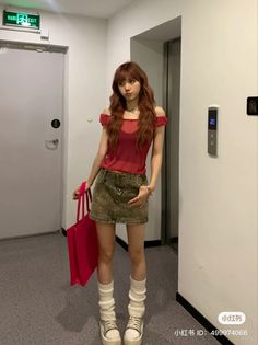 Woman with long hair in a red top and denim skirt holding a pink bag indoors
