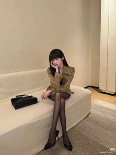 Woman with long black hair wearing a trench coat and black tights sitting on a sofa