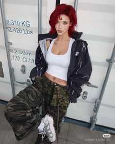 Woman with red hair in a white tank top and camo pants posing in an urban setting