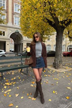 Woman in a denim skirt and brown jacket holding a cup on an autumn street