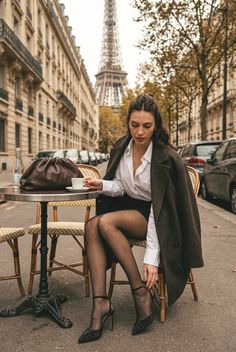 Woman in a brown coat and black tights sitting at an outdoor cafe with the Eiffel Tower in the background
