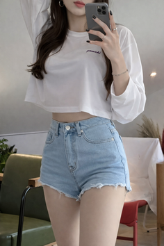 Woman in a white crop top and denim shorts taking a mirror selfie
