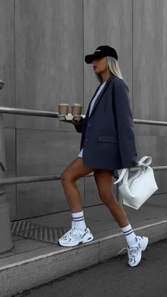 Woman in a blazer and baseball cap carrying coffee and a white bag outdoors