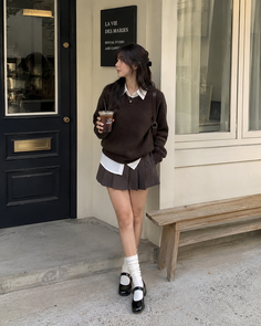 Woman in a brown sweater and pleated skirt holding a drink outdoors