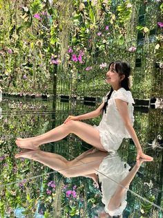 Woman in a white outfit sitting barefoot on a mirrored floor surrounded by lush greenery and pink flowers