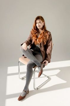 Woman with long red hair in a brown blazer and grey tights sitting on a chair