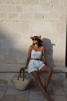 Woman in a straw hat and white shorts sitting outdoors with a straw bag