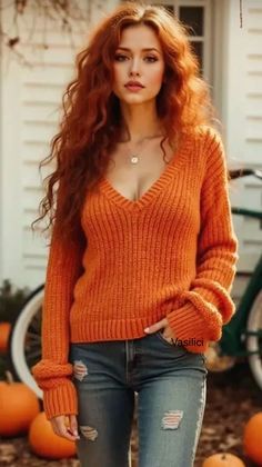 Woman with long red hair in an orange sweater and ripped jeans outdoors with pumpkins