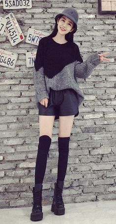 Woman in a bucket hat, oversized sweater, and leather shorts posing against a brick wall