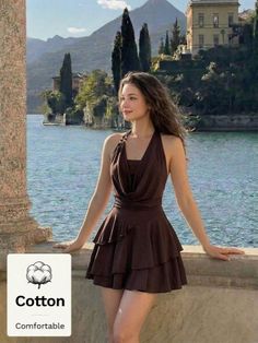 Woman in a brown dress leaning on a stone railing by a lake with mountains
