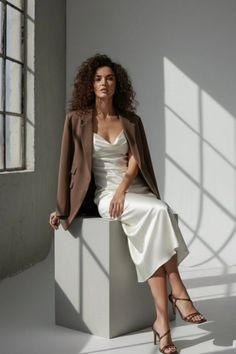 Woman with curly hair in a white slip dress and brown blazer sitting indoors