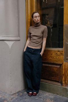 Woman in a brown turtleneck and dark pants leaning against a wall