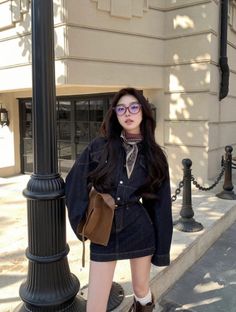 Woman in a denim dress and glasses standing outdoors with a brown bag
