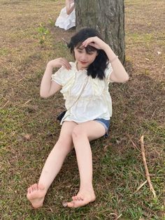 Young woman with black hair sitting barefoot on grass in a ruffled top and denim shorts
