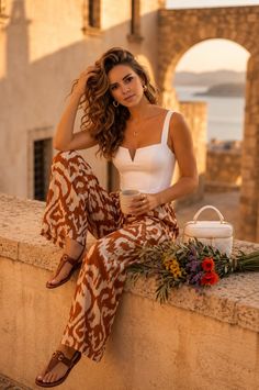 Woman with curly hair in a white top and patterned pants sitting outdoors at golden hour
