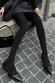 Person in black tights, blazer, and mini skirt sitting on a stone ledge