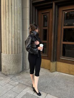 Woman in a black leather jacket and capri pants holding a coffee cup on a city street