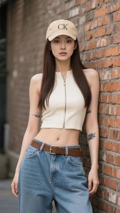 Woman in a crop top, jeans, and baseball cap leaning against a brick wall