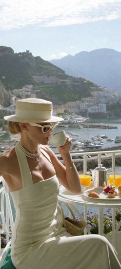 Woman in a straw hat and halter dress having breakfast on a balcony overlooking a coastal town