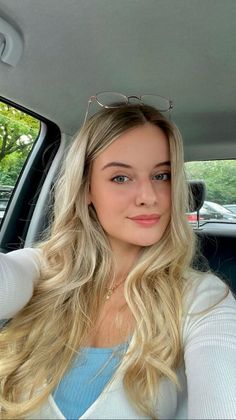 Blonde woman with glasses on her head, wearing a blue top and white cardigan in a car