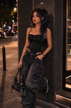 Woman in a black corset and cargo pants leaning against a wall on a city street at night