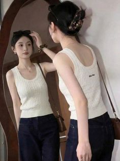 Woman in a white tank top and dark jeans looking at her reflection in a mirror