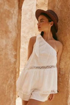 Woman in a white one-shoulder top and straw hat leaning against a wall