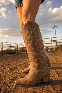 Person wearing cowboy boots and denim shorts on a dirt ground at golden hour
