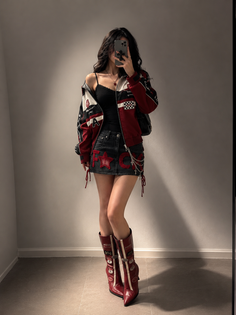 Woman in a racing jacket, denim skirt, and knee-high boots taking a mirror selfie