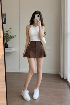 Woman in a white tank top and brown pleated skirt taking a mirror selfie
