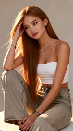Woman with long red hair wearing a white tube top and jeans, sitting with soft lighting